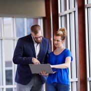 A woman and a man standing near windows looking at a laptop screen.